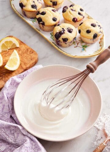 Lemon glaze in a bowl with a whisk. Lemon wedges on a wooden board, a platter of muffins, and a measuring cup with powdered sugar surround it.
