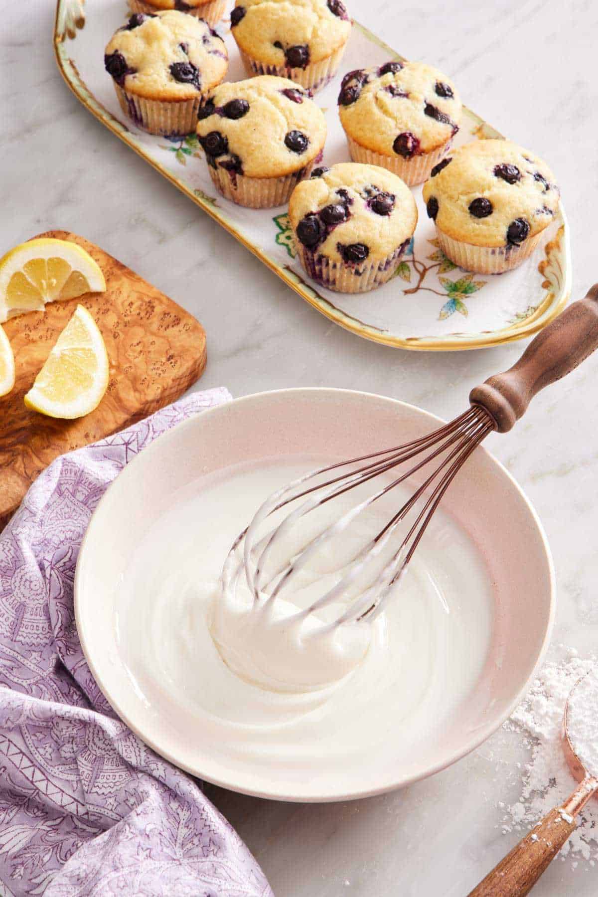Lemon glaze in a bowl with a whisk. Lemon wedges on a wooden board, a platter of muffins, and a measuring cup with powdered sugar surround it.