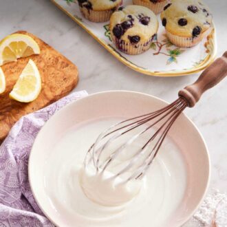 Pinterest graphic showing glaze in a bowl surrounded by lemon wedges, homemade muffins on a platter, and powdered sugar in a measuring cup.