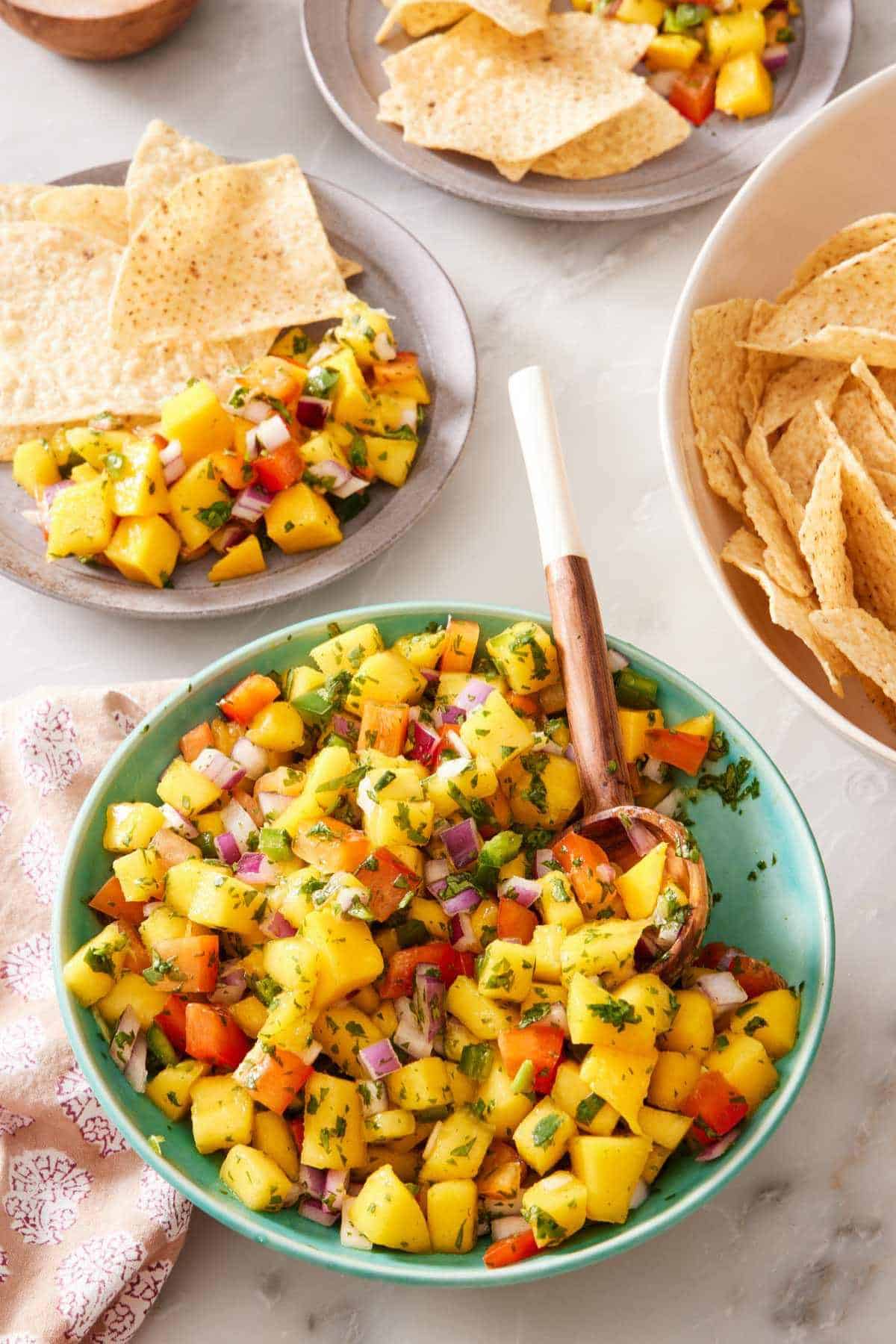 Mango salsa in a bowl with a wooden spoon, with servings of salsa and tortilla chips in the background.