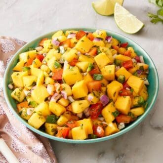 Mango salsa in a bowl with a wooden spoon, limes, cilantro, and salt on the side.