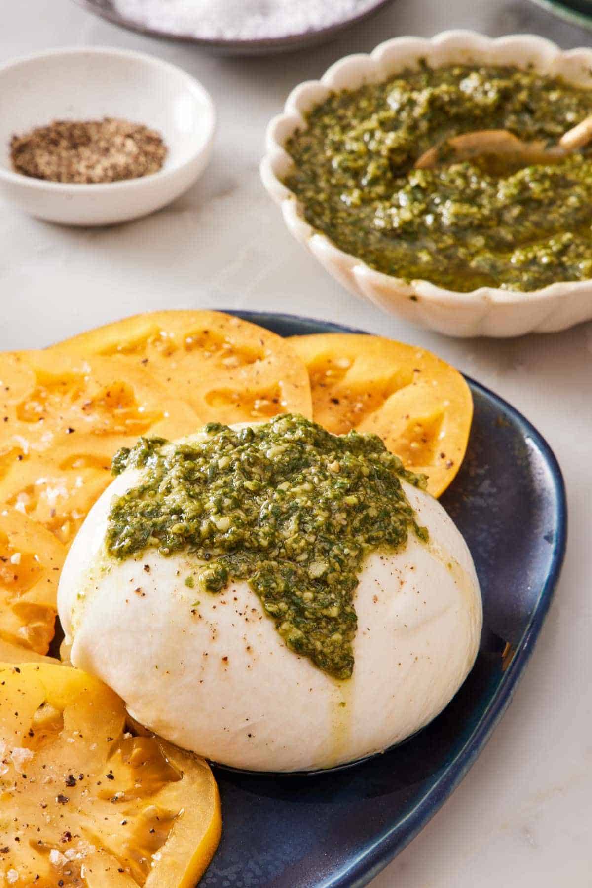 Burrata topped with basil pesto with sliced tomatoes on the side. A bowl of the sauce is in the background.