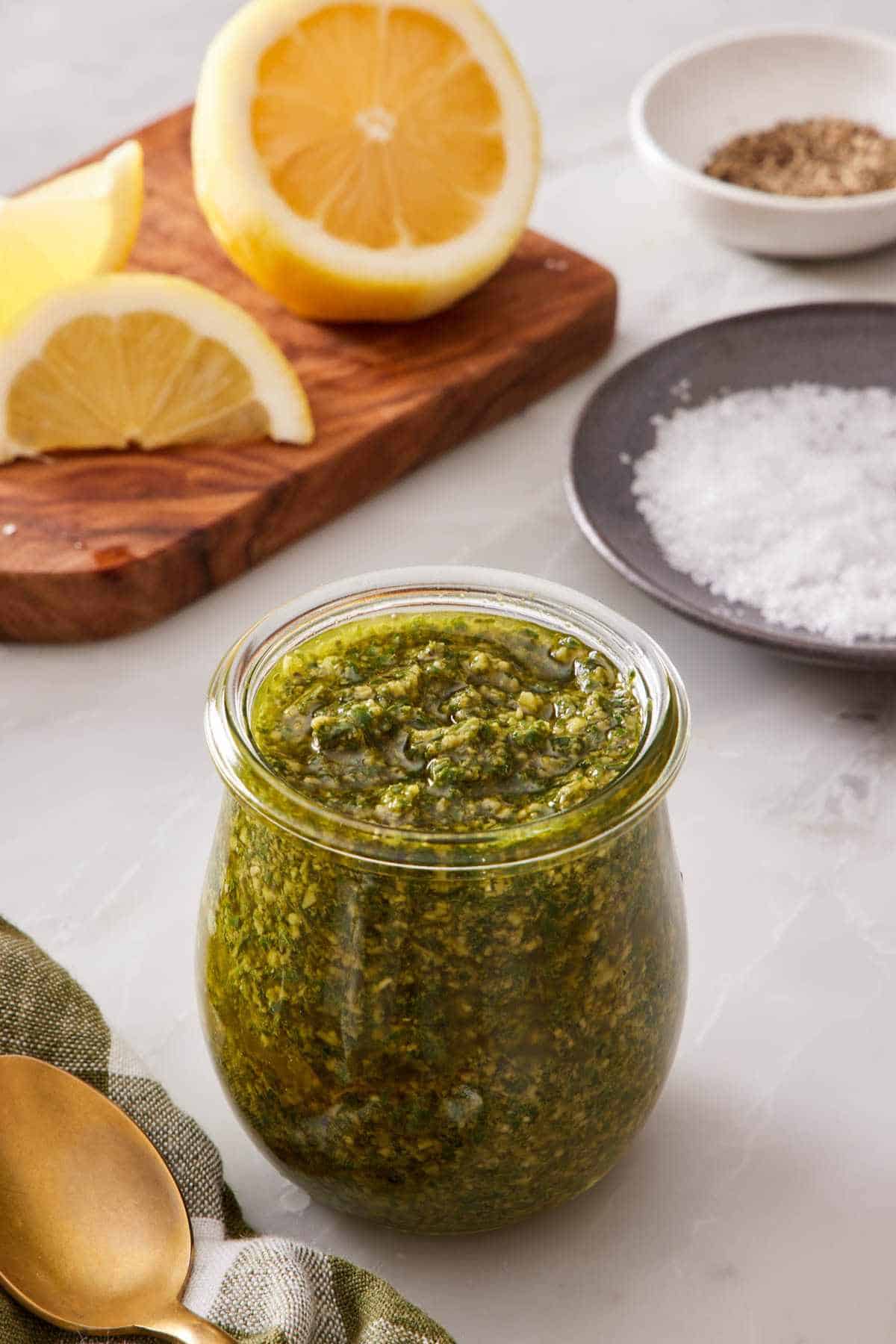 A small glass jar of homemade pesto sauce with lemon, salt, and pepper in the background.