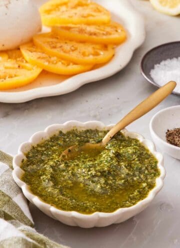 Pesto in a white bowl with a spoon, with sliced tomatoes in the background.