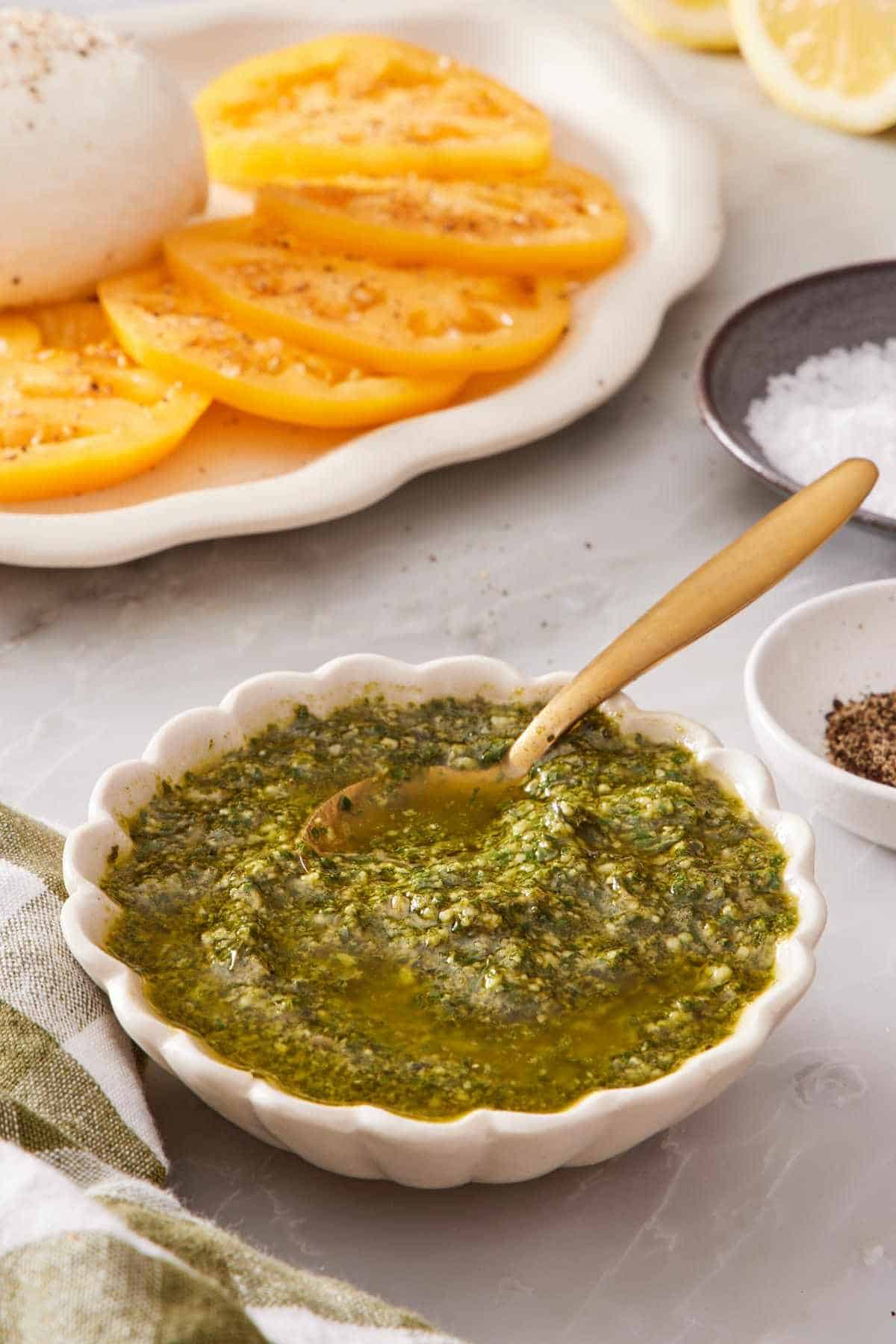 Pesto in a white bowl with a spoon, with sliced tomatoes in the background.