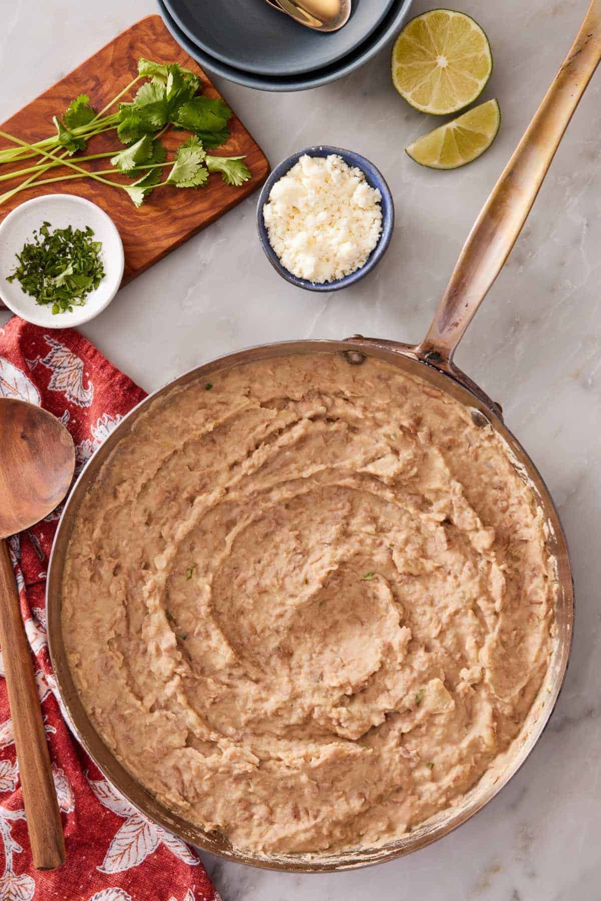 Overhead shot of refried beans in a large skillet with queso fresco, limes, and cilantro on the side.