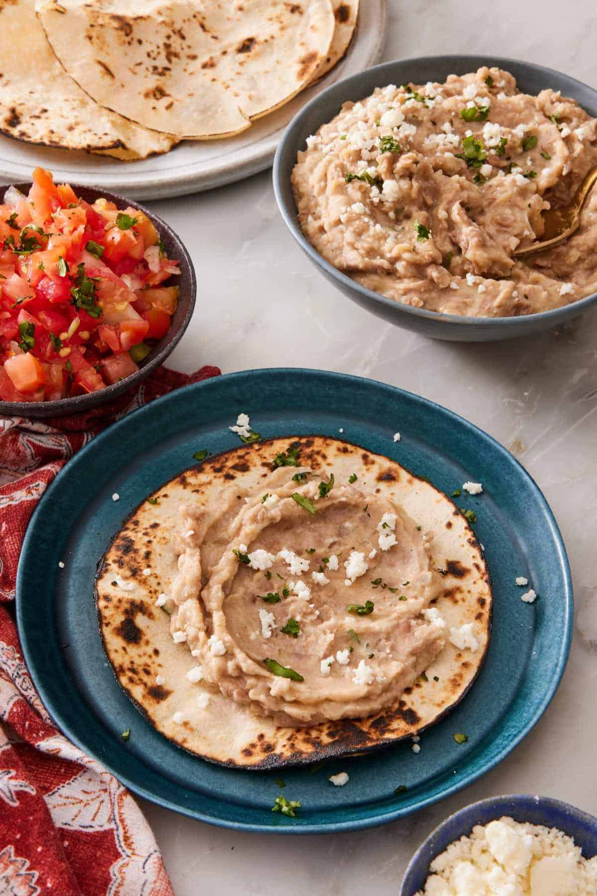 Refried beans spread on a toasted tortilla with more beans, pico de gallo, and more tortillas in the background.