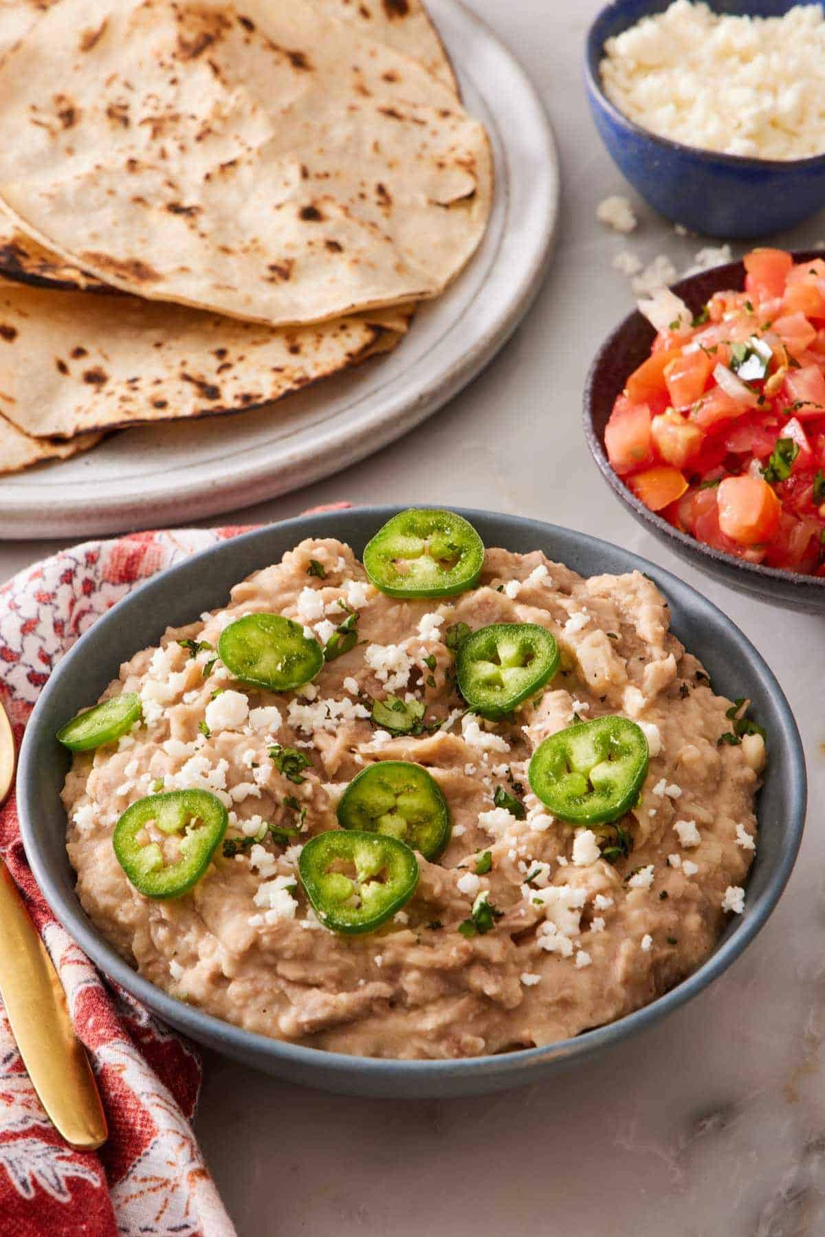 Homemade refried beans topped with queso fresco, cilantro, and jalapenos. Pico de gallo and tortillas in the background.