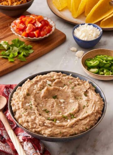 Mashed pinto beans in a bowl with pico de gallo, jalapenos, queso fresco, and hard tortilla shells in the background.