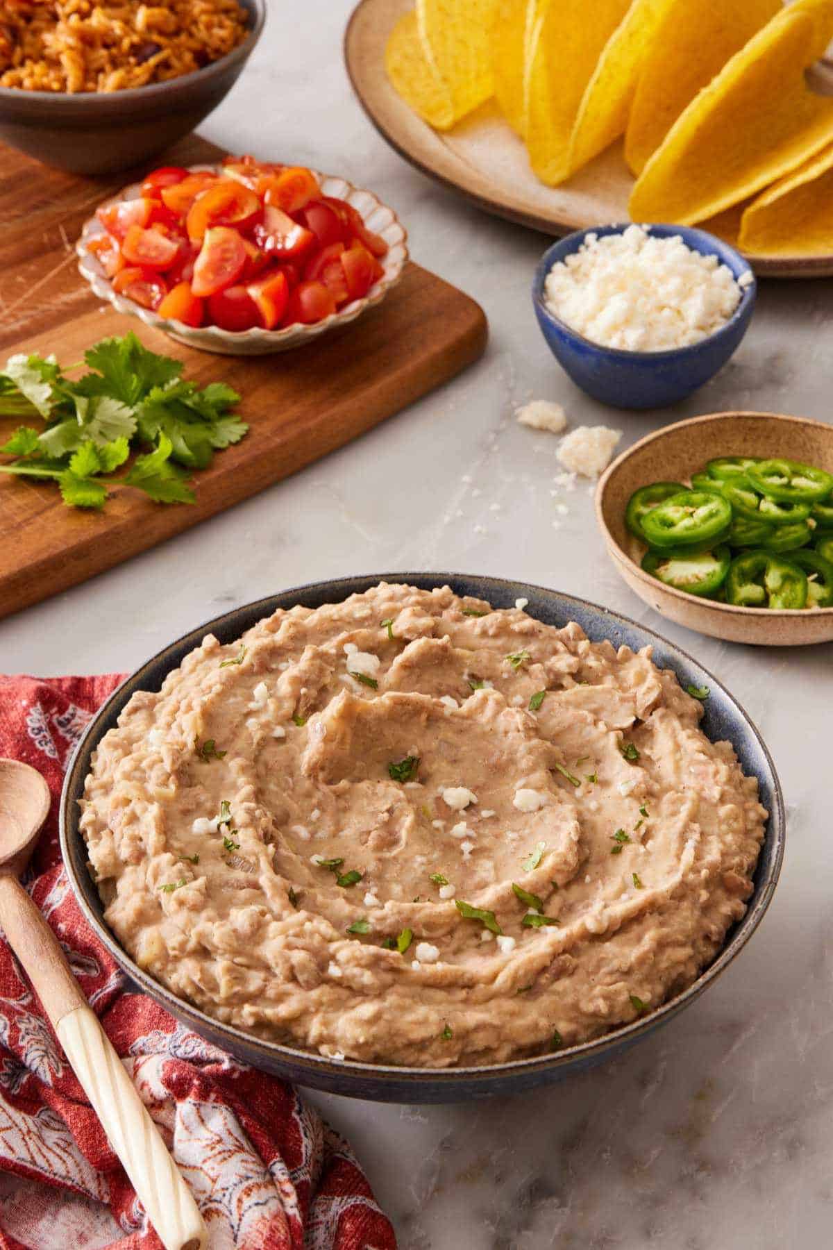 Mashed pinto beans in a bowl with pico de gallo, jalapenos, queso fresco, and hard tortilla shells in the background.
