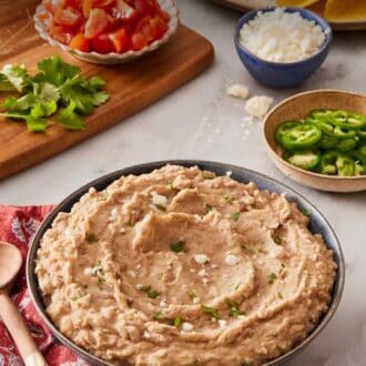 Pinterest graphic showing refried beans in a large bowl with toppings like jalapenos, cheese, and diced tomato in the background.