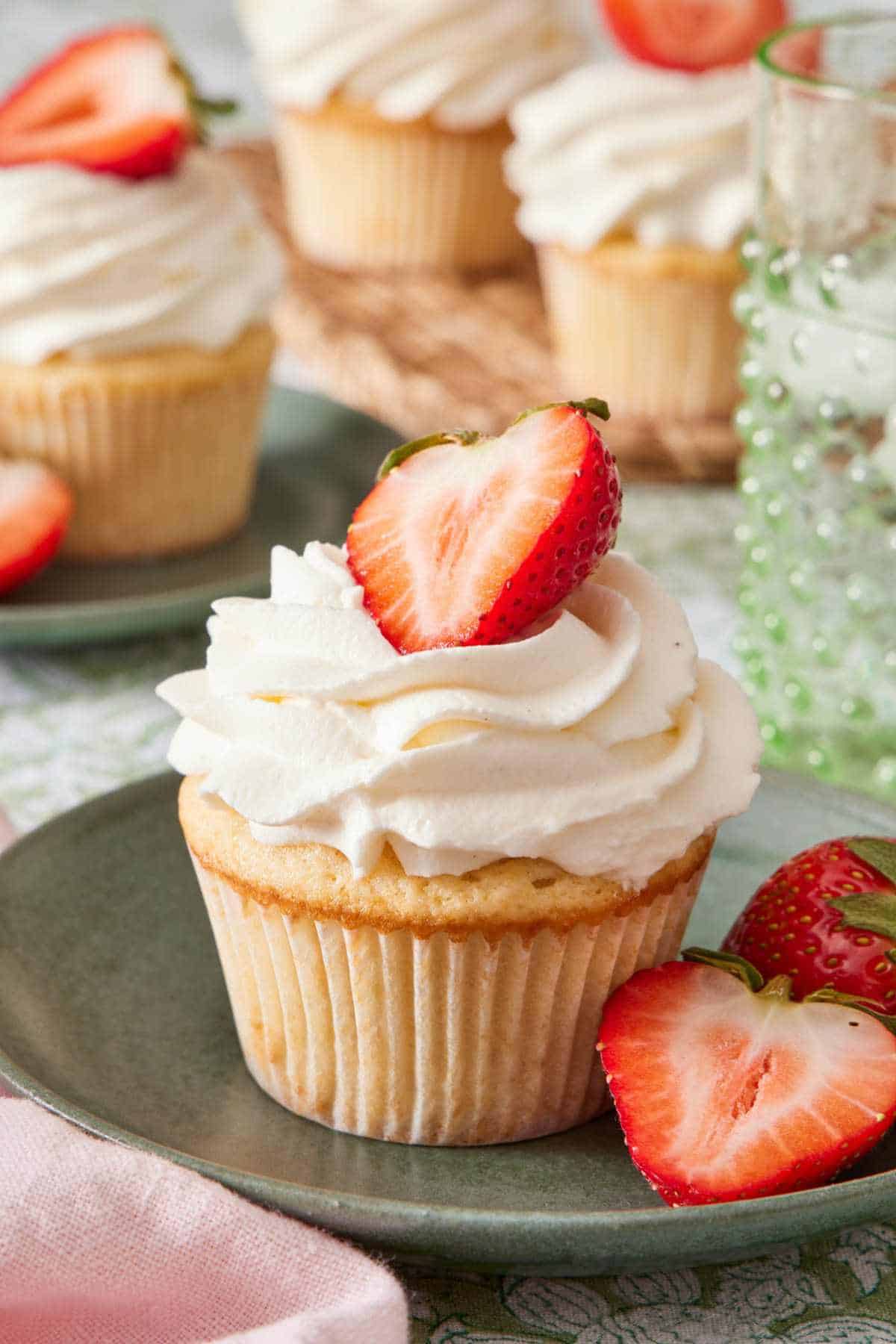 Strawberry shortcake cupcake on a plate with whipped topping and strawberry halves. More cupcakes in the background.