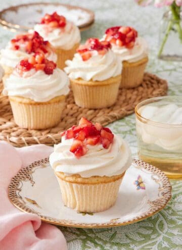 Cupcake on a plate topped with whipped topping and diced strawberries. A drink sits alongside it, and there's more strawberry shortcake cupcakes in the background.
