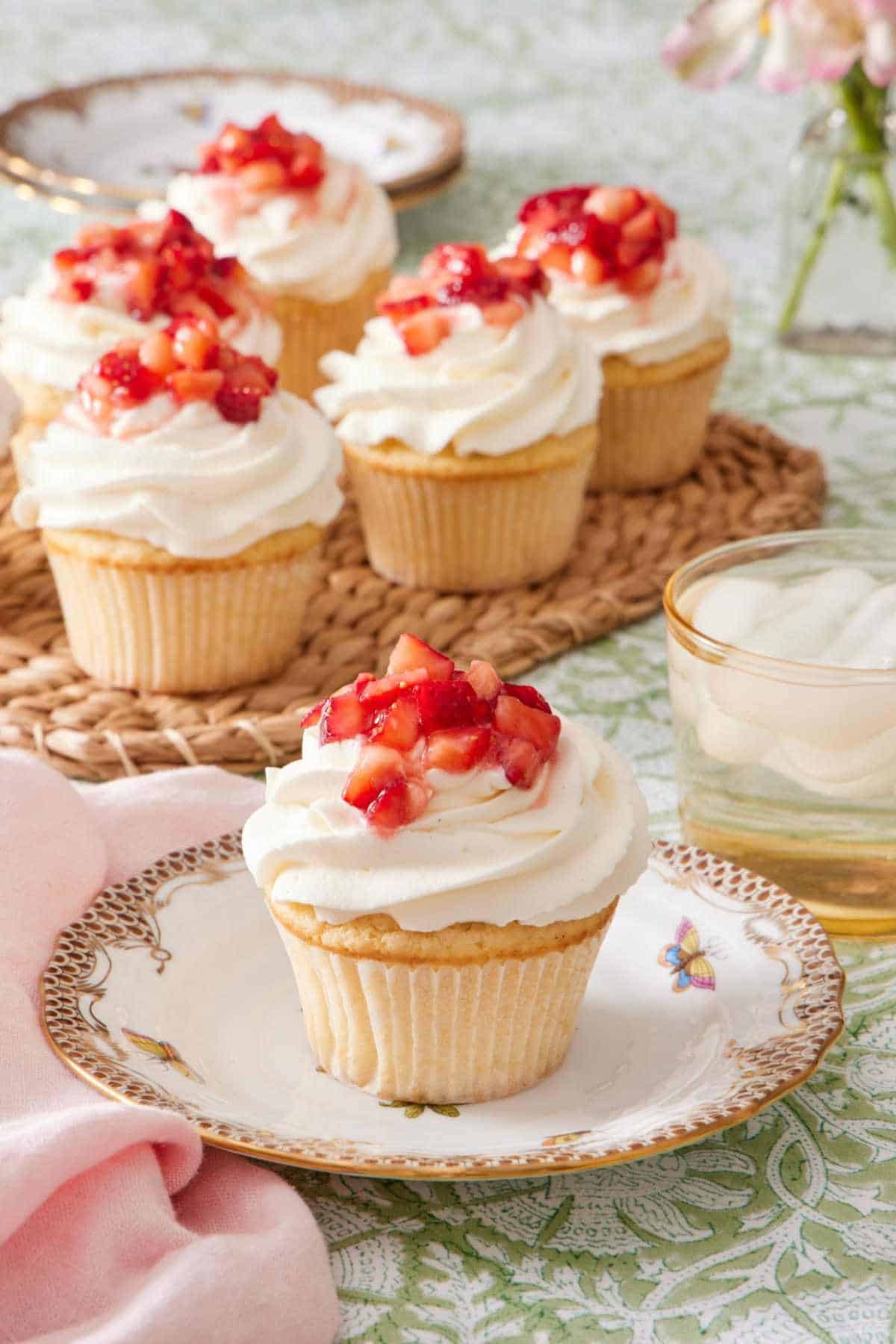 Cupcake on a plate topped with whipped topping and diced strawberries. A drink sits alongside it, and there's more strawberry shortcake cupcakes in the background.
