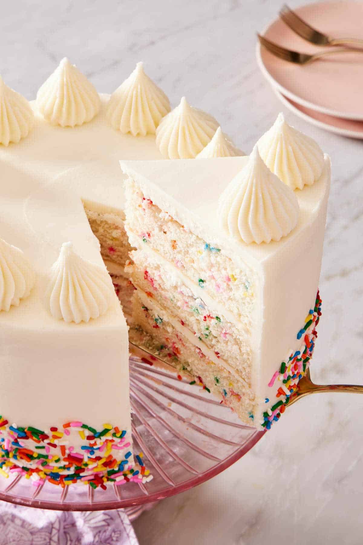 A slice of confetti cake on a cake server next to the rest of the cake showcasing the colorful layers with sprinkles.