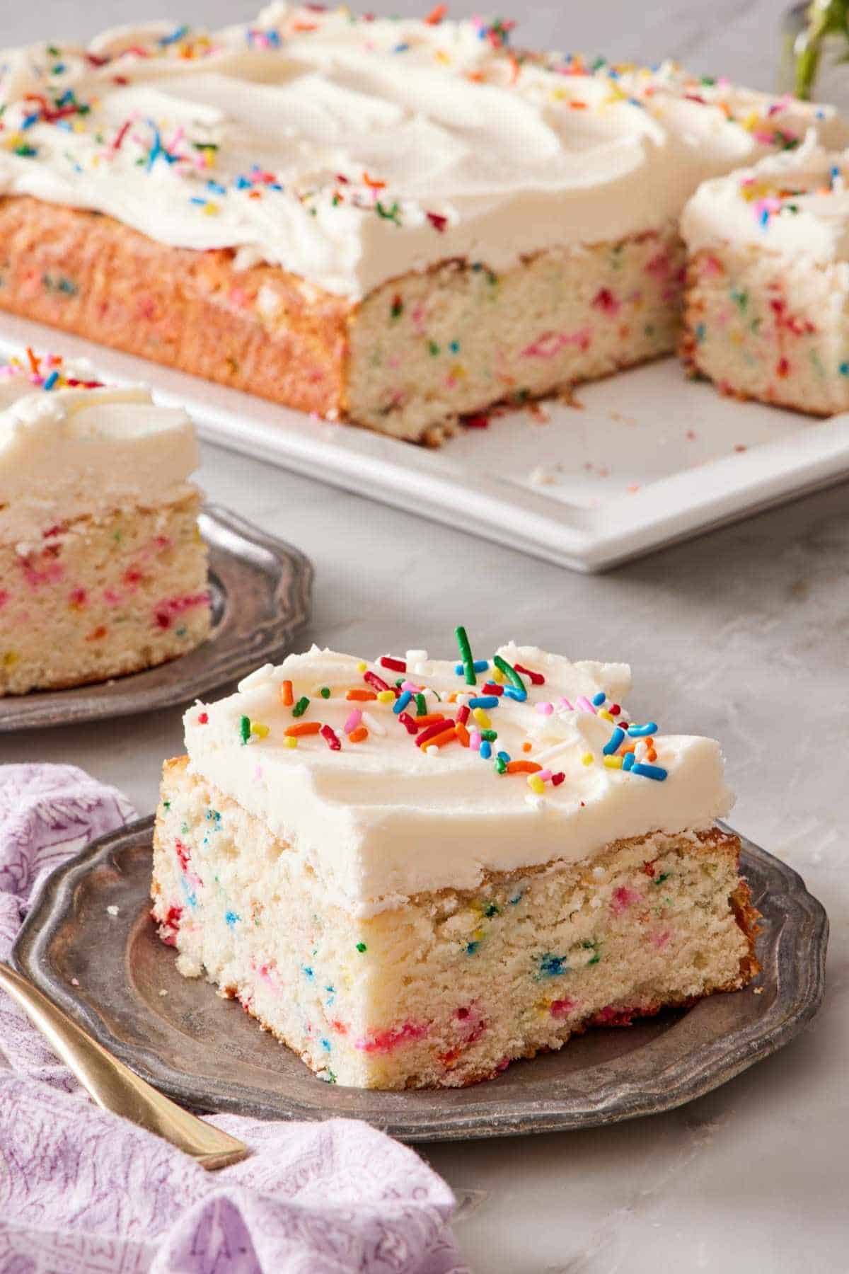 Slice of funfetti sheet cake with buttercream frosting on a small plate. The rest of the cake in the background.