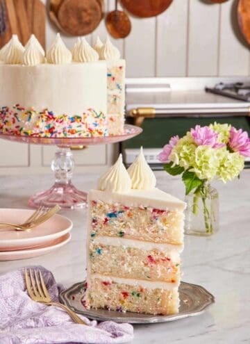 A slice of three-layer funfetti cake on a small plate with a vase of flowers right behind. The rest of the cake in the background.