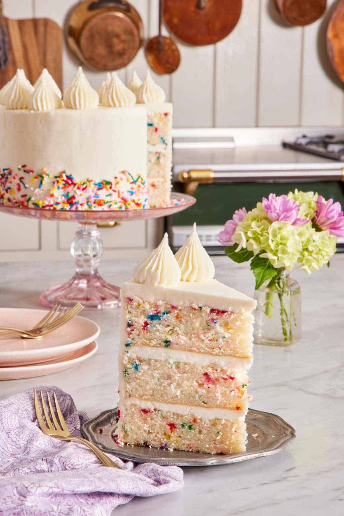 A slice of three-layer funfetti cake on a small plate with a vase of flowers right behind. The rest of the cake in the background.