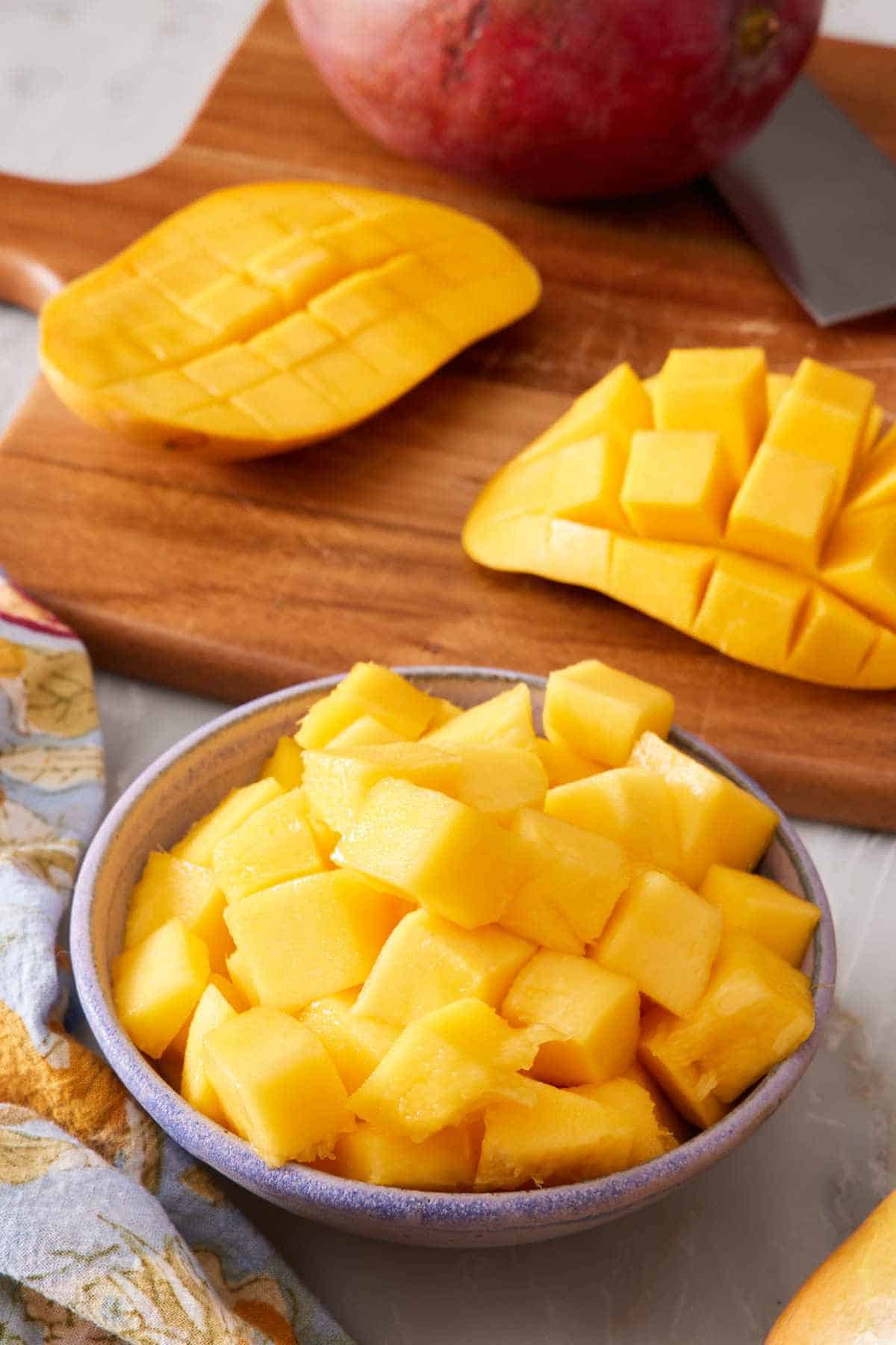 Cubed fresh mango in a small bowl with scored mango halves in the background on a wooden cutting board.