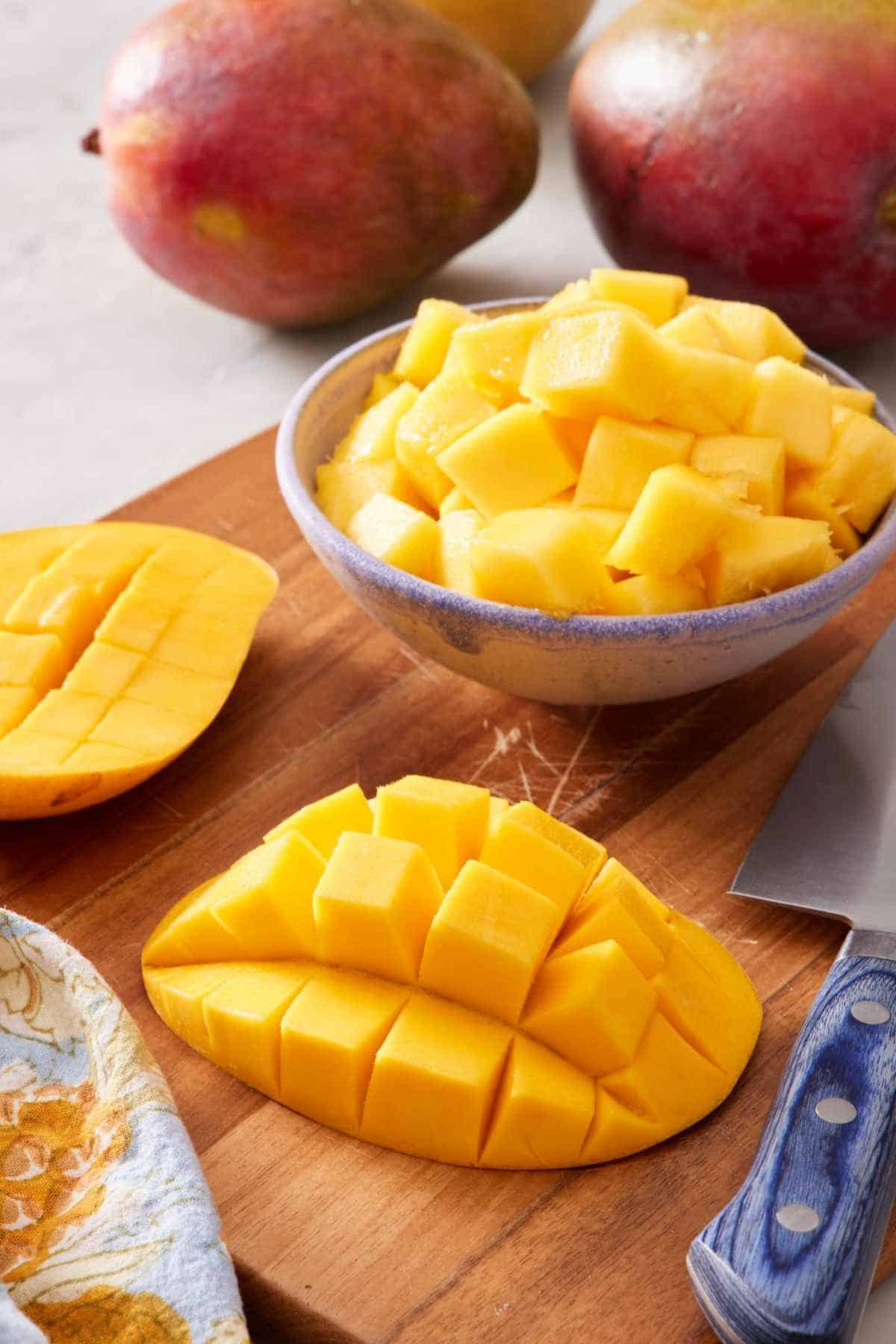 Mango cheeks with a grid cut into them, and cubed mango in a bowl. Whole mangoes in the background.