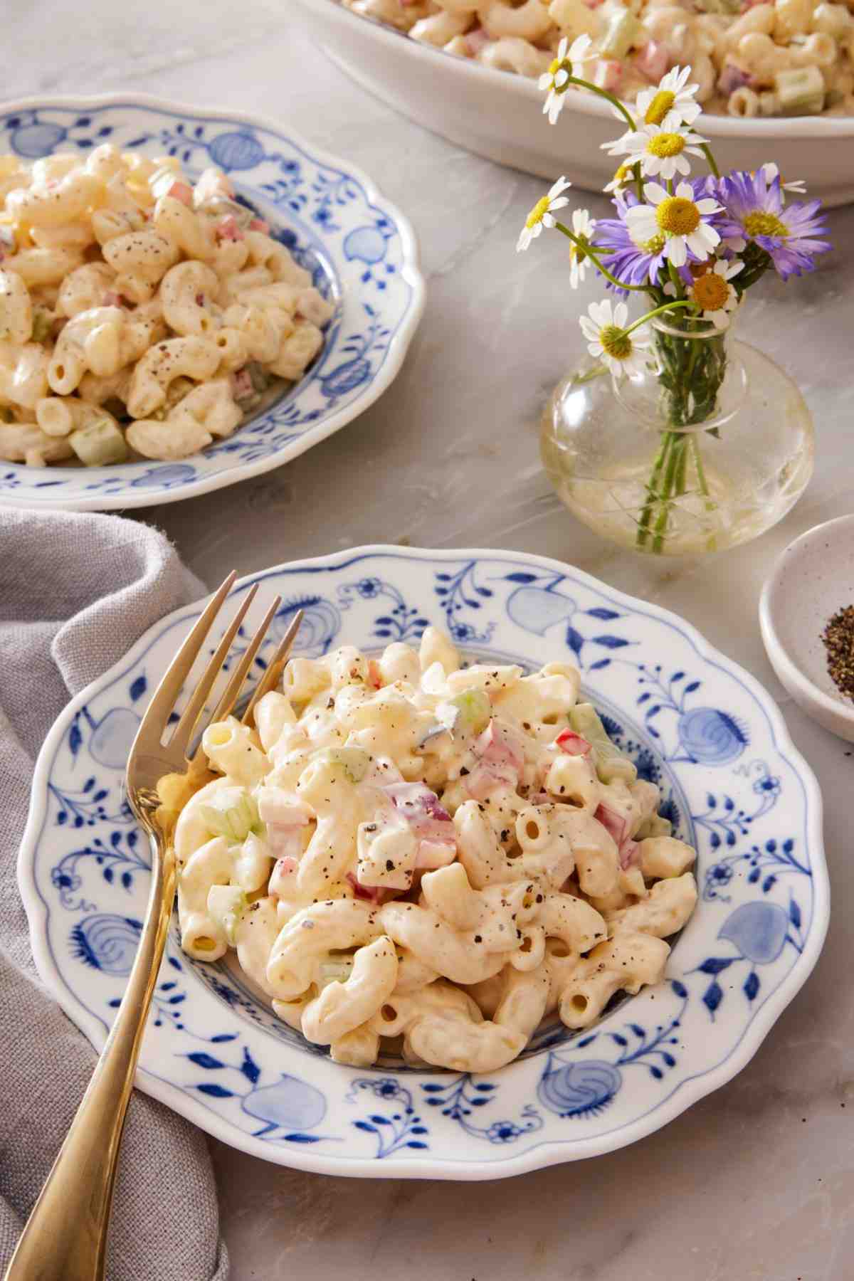 Creamy macaroni salad in a blue and white bowl with a gold fork. A small vase of flowers alongside.