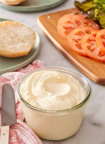 A small glass jar of mayo with sliced tomatoes and greens in the background.