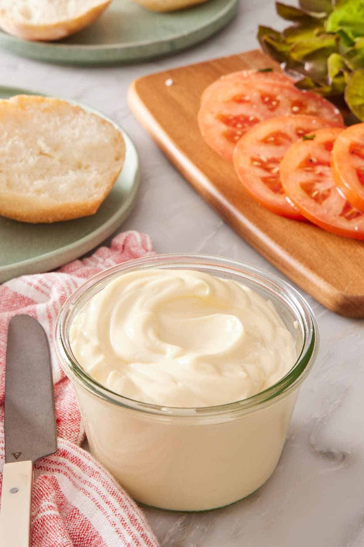 A small glass jar of mayo with sliced tomatoes and greens in the background.