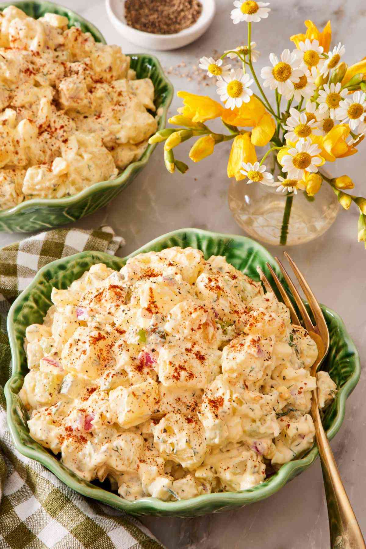 Classic potato salad sprinkled with paprika in a green bowl with a small vase of flowers next to it.