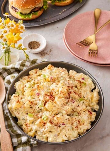 A large bowl of creamy salad with plates and burgers in the background.