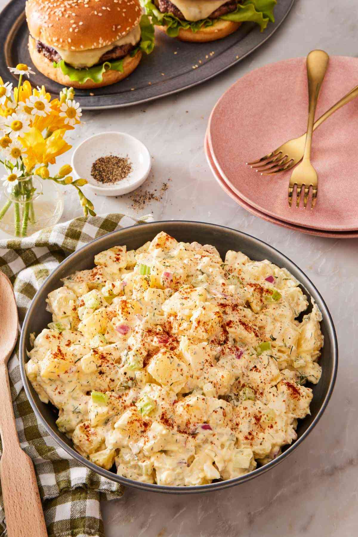 A large bowl of creamy salad with plates and burgers in the background.