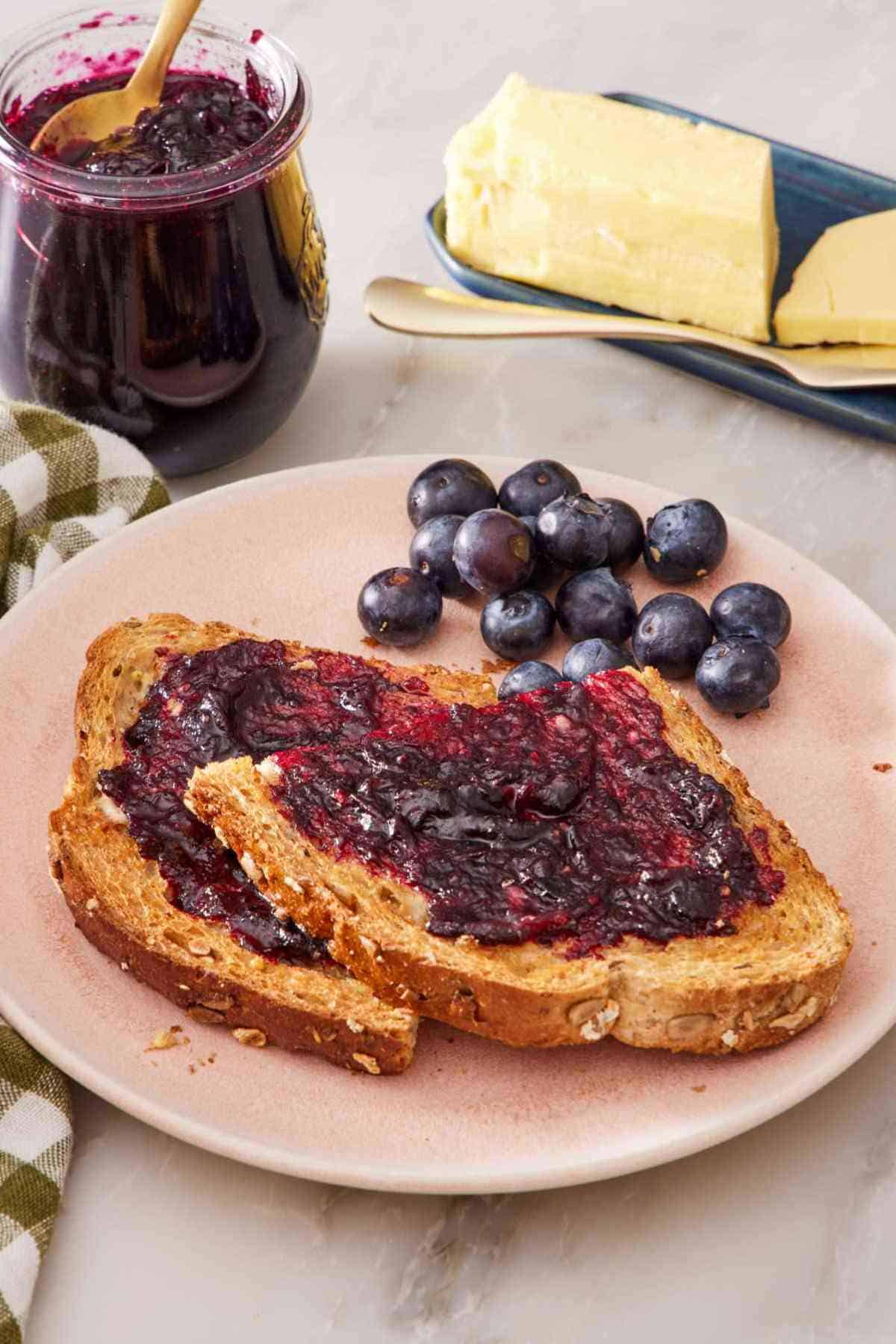 A plate with fresh blueberries and two slices of toast with a smear of jam on both. A stick of butter and a small jar of jam in the background.