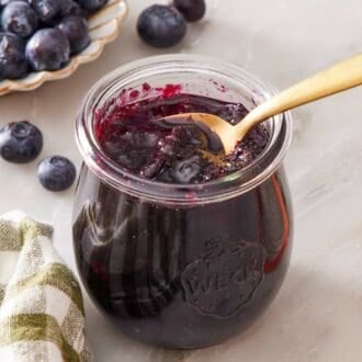 Close up of a small glass jar of homemade jam with a spoon. A green and white linen sits alongside.