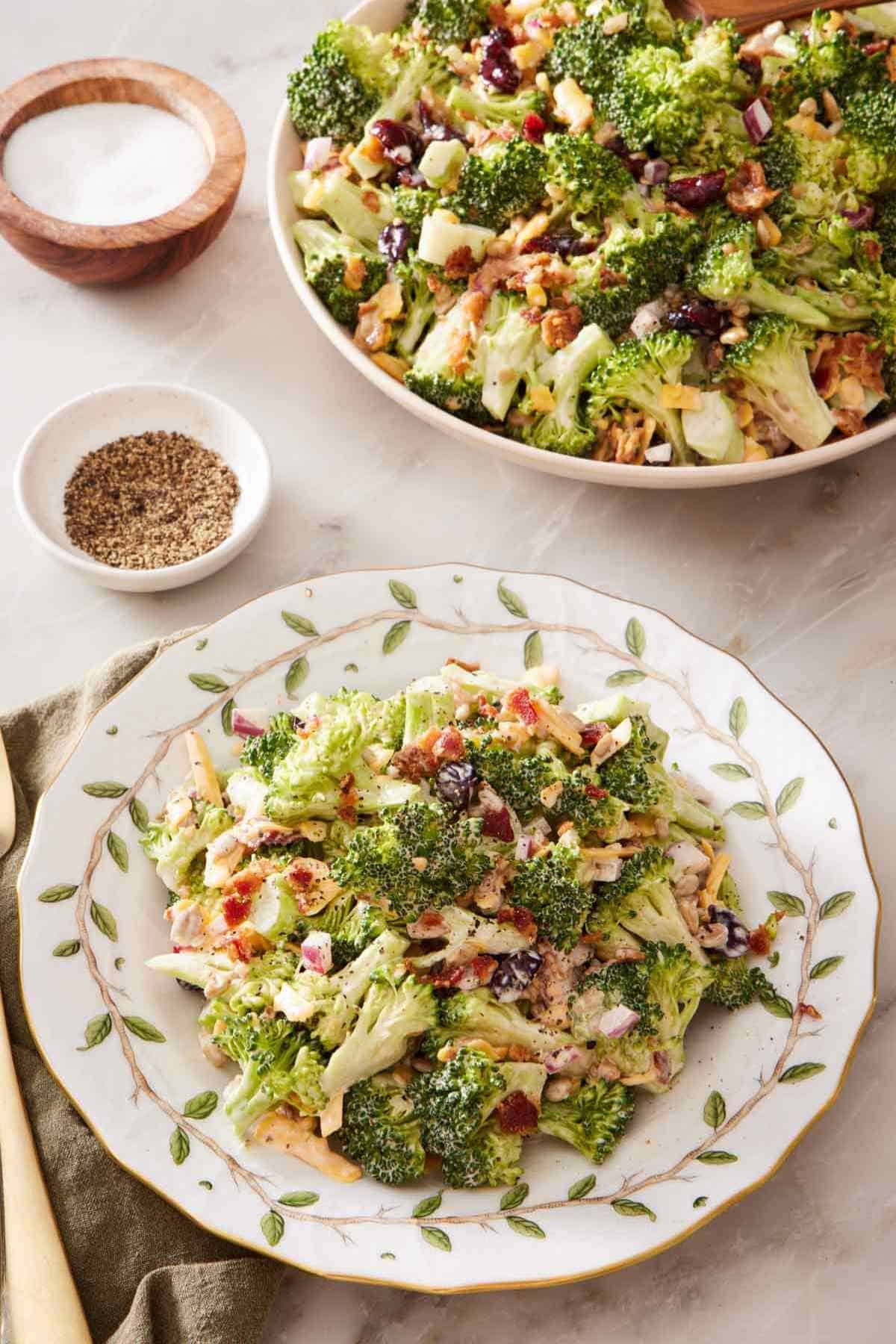 Broccoli salad with a white plate patterned with leaves. Salt and pepper alongside, and a big bowl of broccoli salad in the background.