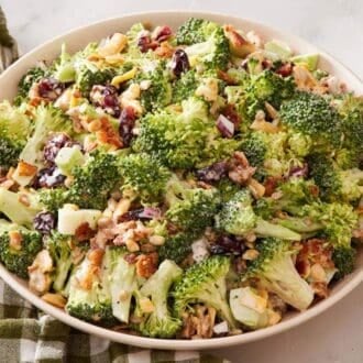 Broccoli salad in a large white bowl with a wooden spoon resting on a table linen on one side, and a small bowl of black pepper on the other.