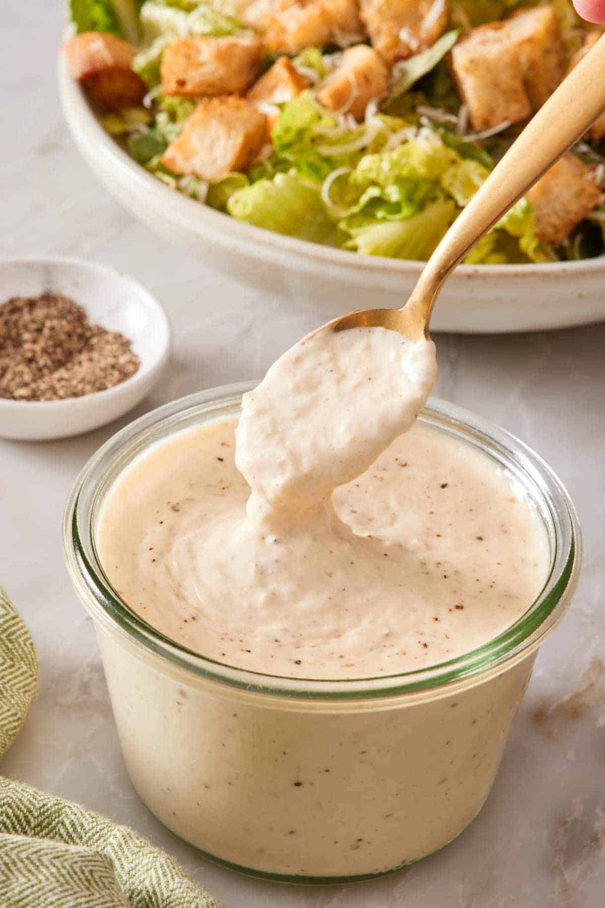 A spoon dipped into a jar of homemade Caesar dressing with an undressed Caesar salad in the background.