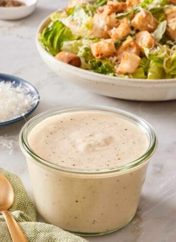 A small jar of Caesar dressing with a salad in the background.