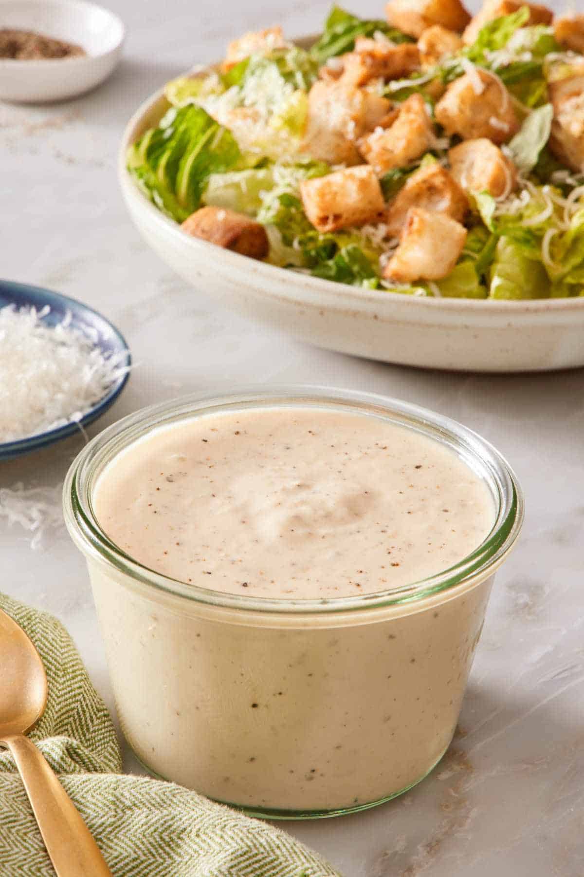 A small jar of Caesar dressing with a salad in the background.