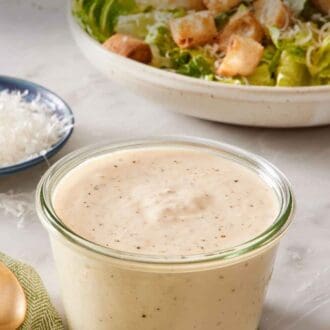 Pin image showing a small jar of creamy Caesar salad dressing with a Caesar salad in the background.