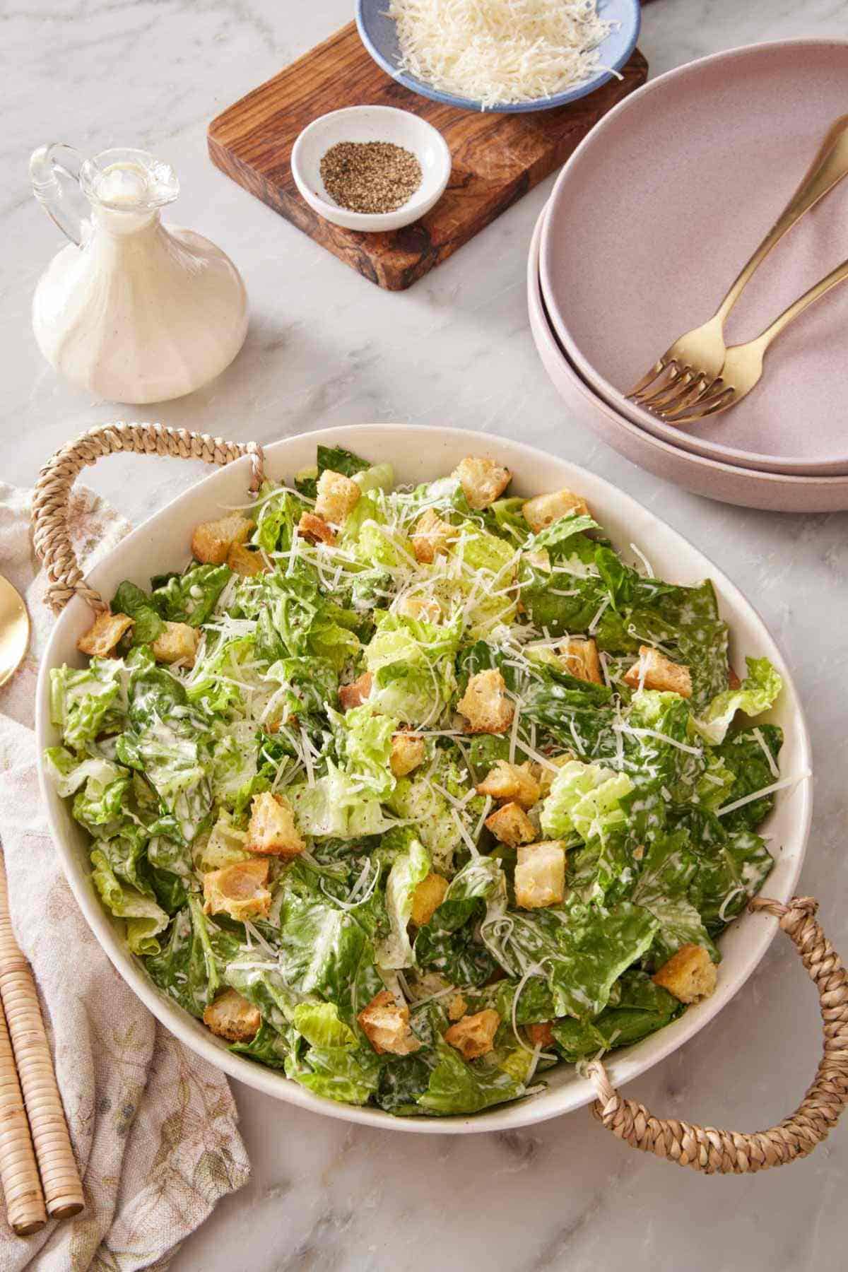 A serving bowl with Caesar salad with plates, salad dressing, pepper, and parmesan cheese on the side.