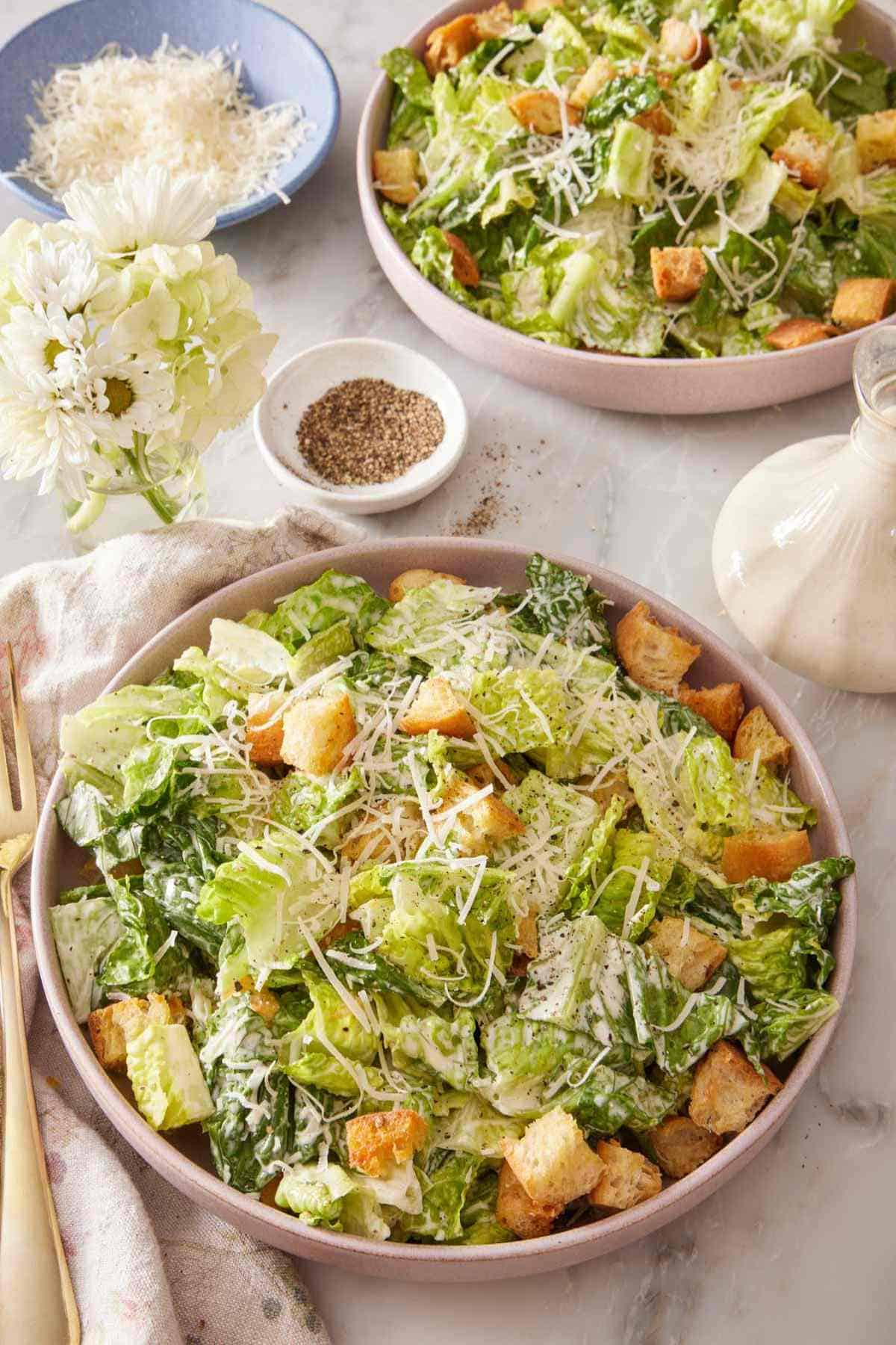Two plates of Caesar salad with salad dressing, cheese, and black pepper on the side.