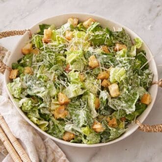 Caesar salad in a white serving bowl with serving utensils on a linen on the side.