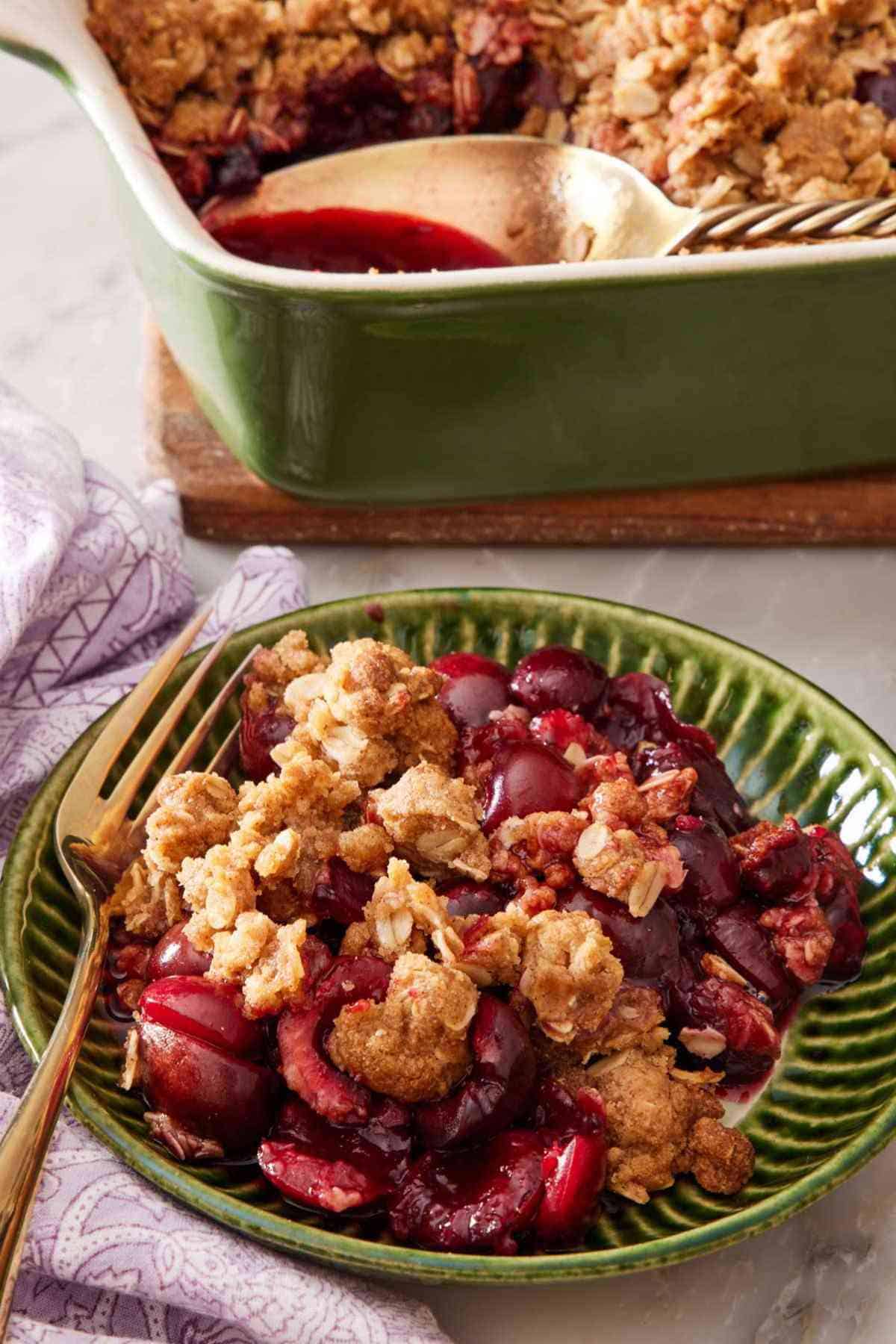 Homemade cherry crisp in a textured green bowl with a casserole dish containing the rest of the dessert in the background.