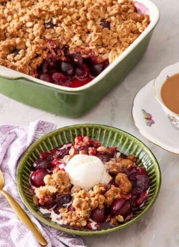 Cherry crisp in a green bowl with a scoop of vanilla ice cream on top. A cup of tea and a baking dish containing the rest of the crisp is in the background.