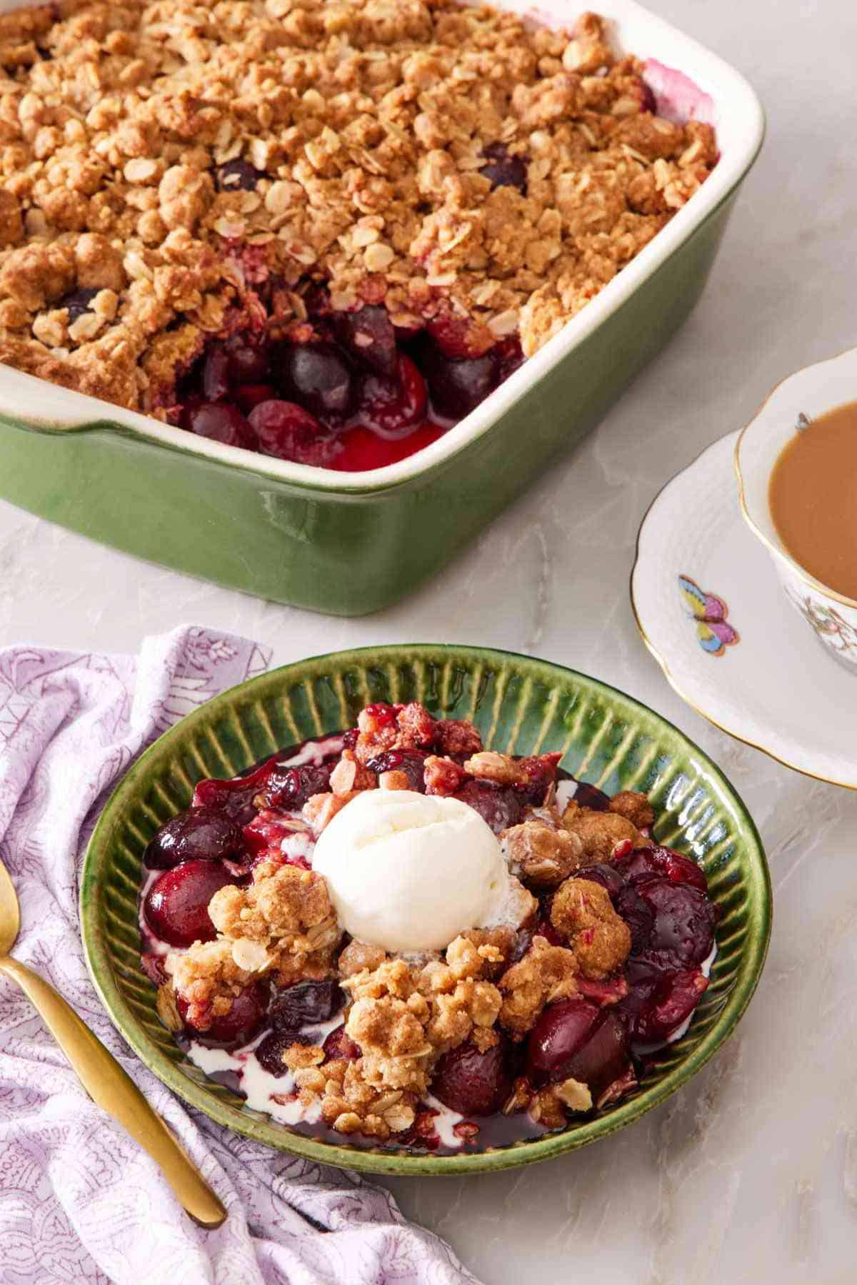 Cherry crisp in a green bowl with a scoop of vanilla ice cream on top. A cup of tea and a baking dish containing the rest of the crisp is in the background.