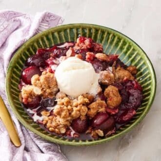 Cherry crisp in a green bowl with a scoop of ice cream on top. A golden spoon and lilac table linen is next to the bowl.