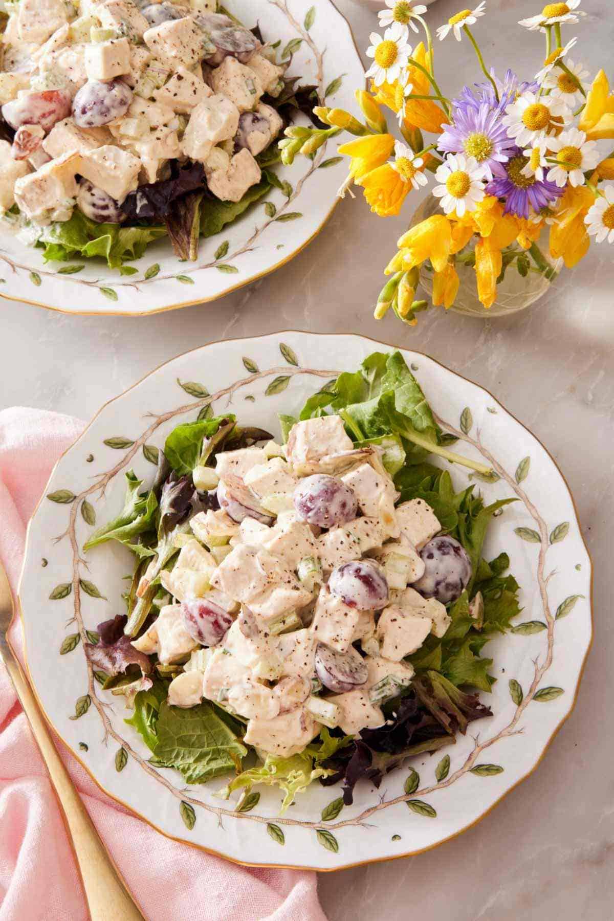 Two plates of chicken salad served over a bed of greens with a vase of flowers next to them.