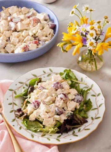 Chicken salad on mixed greens on a patterned plate with a vase of flowers and more salad alongside.