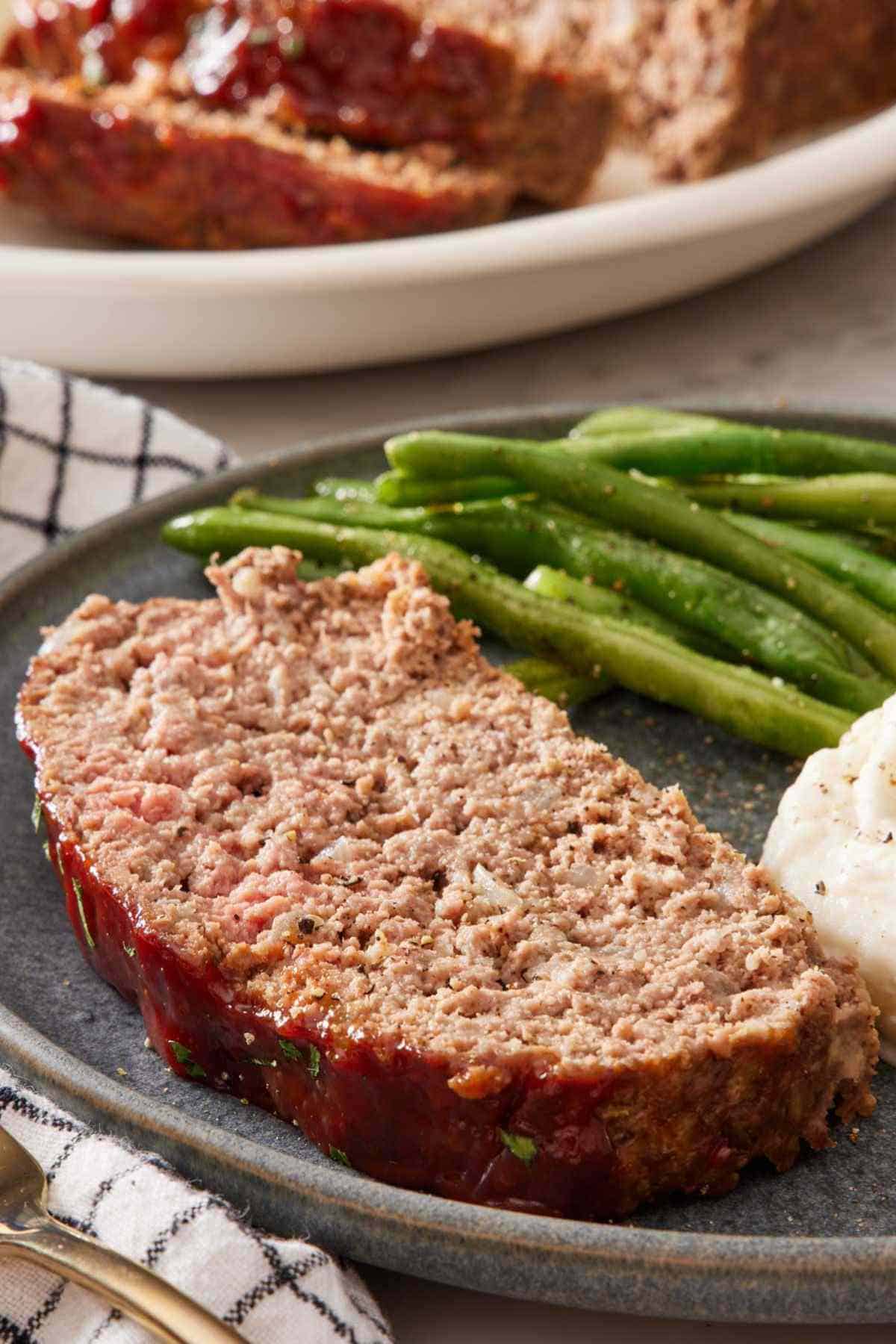 A slice of meatloaf on a plate with green beans and mashed potatoes.