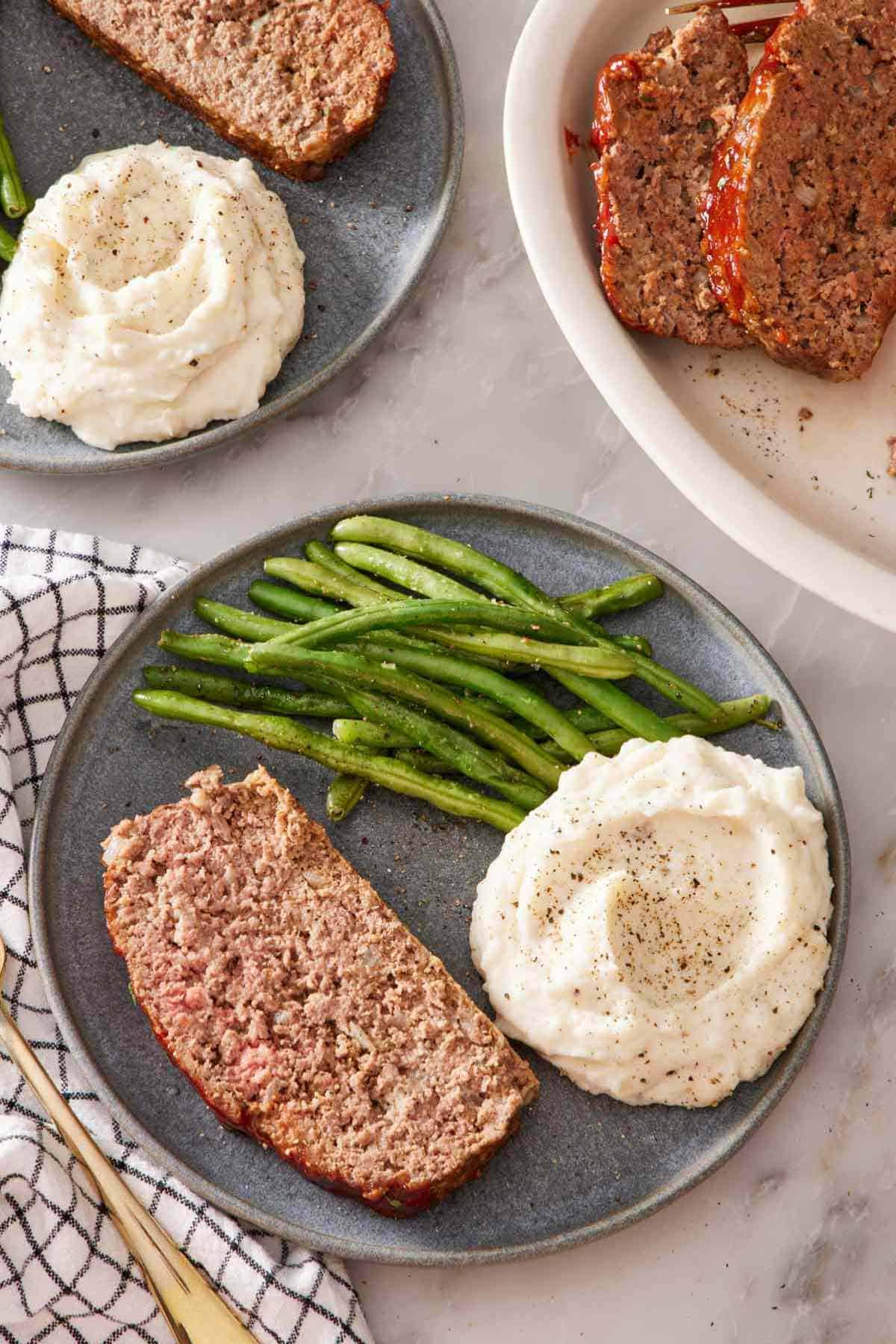 Overhead shot of a plate with a slice of meatloaf, green beans, and mashed potatoes. Another plated meal is off to the side, and a dish of sliced meatloaf is next to it.