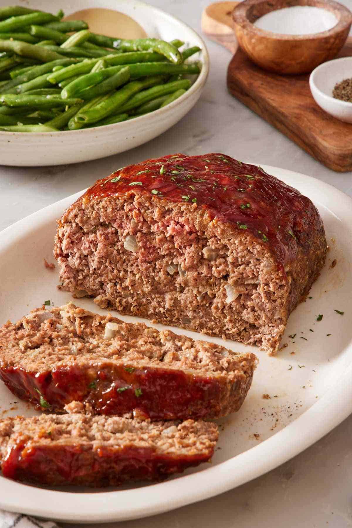 Meatloaf with two slices cut and laying on a serving platter. Dish of green beans in the background.